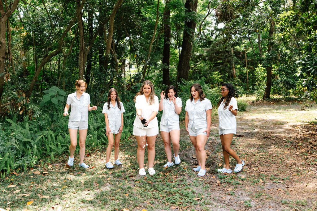 The bride and her bridesmaids celebrate outdoors in matching pajamas and Crocs as the bride pops a bottle of champagne. Surrounded by lush greenery, the group shares a candid and joyful moment during the wedding morning festivities.