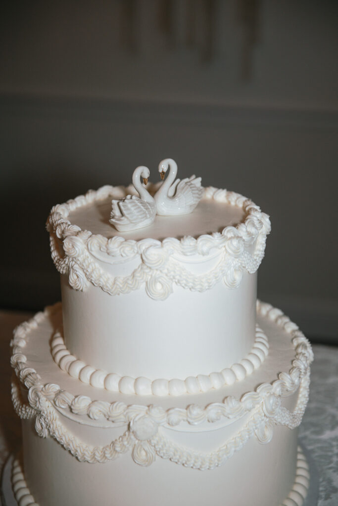 Vintage-inspired white wedding cake with intricate piping and porcelain swan toppers, perfect for a traditional wedding celebration.