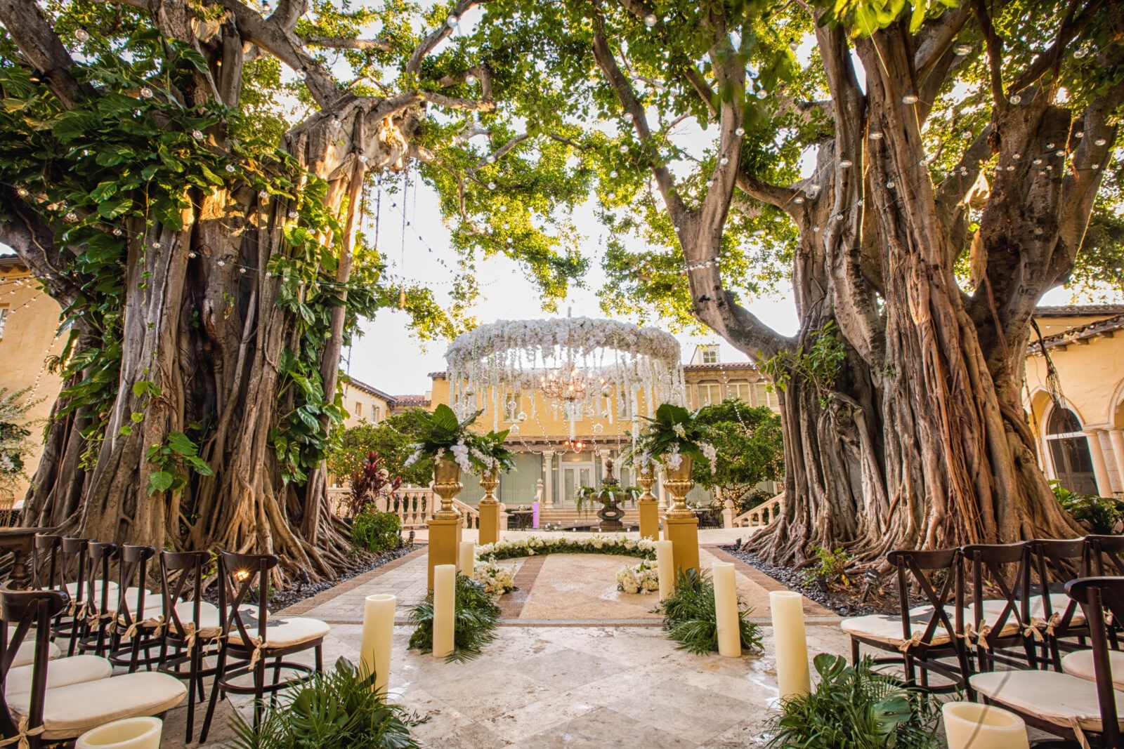 A breathtaking outdoor wedding ceremony space framed by towering banyan trees. The aisle is lined with white petals, large candles, and lush greenery, leading to a romantic floral chandelier altar under a natural canopy of branches.